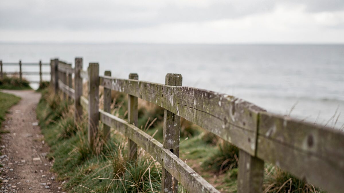 A weathered wooden fence running along an English coastal path under soft overcast light, photographed from a low angle with the grey sea blurred behind.
